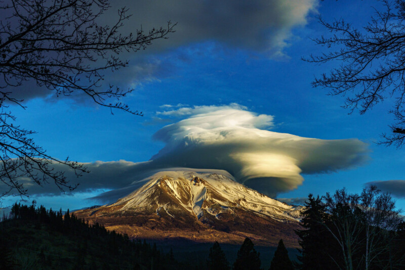 Snow-capped mountain with dramatic, layered lenticular clouds above it, surrounded by trees and branches silhouetted against a vibrant blue and golden sky.