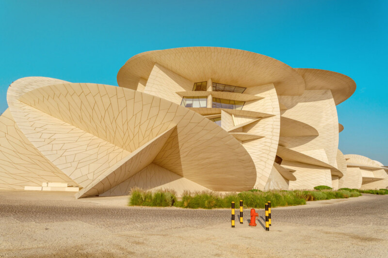 A modern beige building with large, overlapping, disk-shaped layers, resembling desert rose crystals, under a clear blue sky. Two yellow-black posts and a red hydrant are in the foreground.