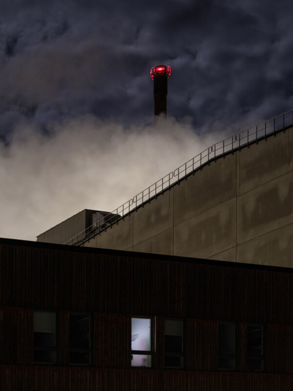 A tall smokestack with a red warning light rises above an industrial building at night, emitting wisps of smoke that blend into a dark, cloudy sky. The building has lit windows and metal railings along the roof.