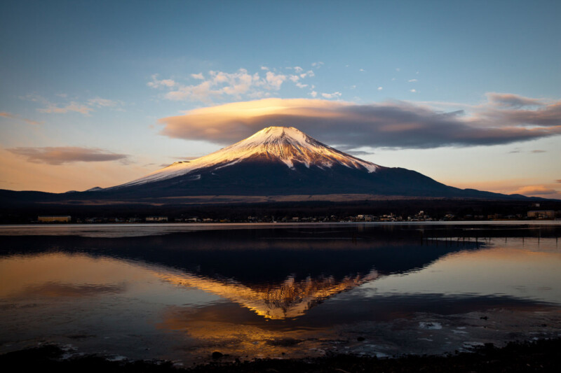 Snow-capped Mount Fuji at sunrise, with clouds above and its reflection visible in a calm lake below. The sky is mostly clear with soft, warm lighting.