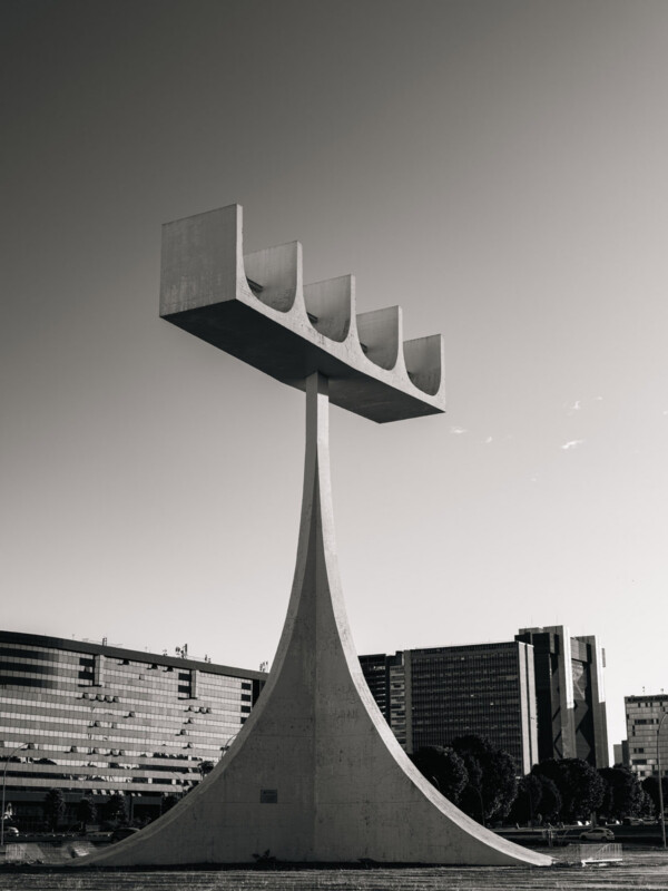 A tall concrete monument with a curved base and four rectangular prongs at the top, set against a cityscape with modern buildings in the background, photographed in black and white.