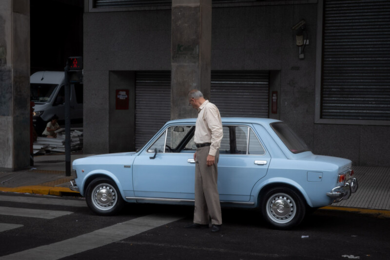 An older man in a light shirt and beige pants stands beside a light blue vintage car parked at a curb in an urban area with gray buildings and closed metal shutters in the background.