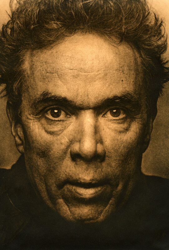 A sepia-toned close-up portrait of an older man with intense eyes, deep-set wrinkles, and wavy hair. His expression is serious and direct, with dramatic lighting highlighting his facial features.