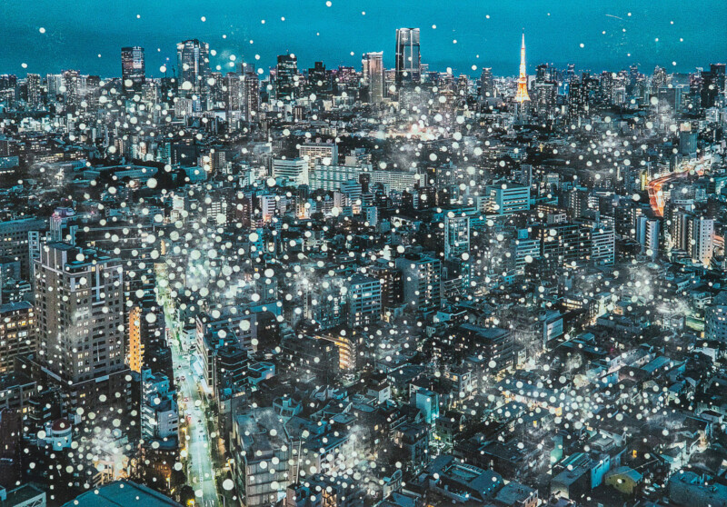 A vibrant cityscape at night with numerous tall buildings and skyscrapers illuminated by lights. White dots are scattered across the image, giving a dreamy, sparkling effect. The sky is a deep blue.