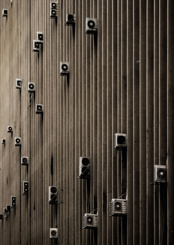 Rows of air conditioning units are mounted on the exterior of a tall, vertically-striped building wall, creating a geometric and repetitive pattern in a sepia-toned image.
