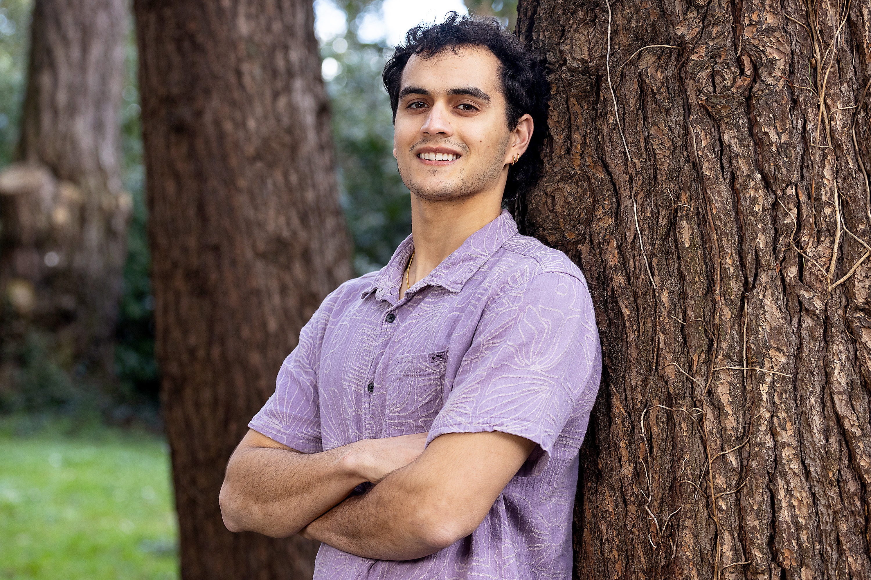 Dillon Chana, a 23-year-old student, stands next to a tree with his arms crossed, smiling at the camera.