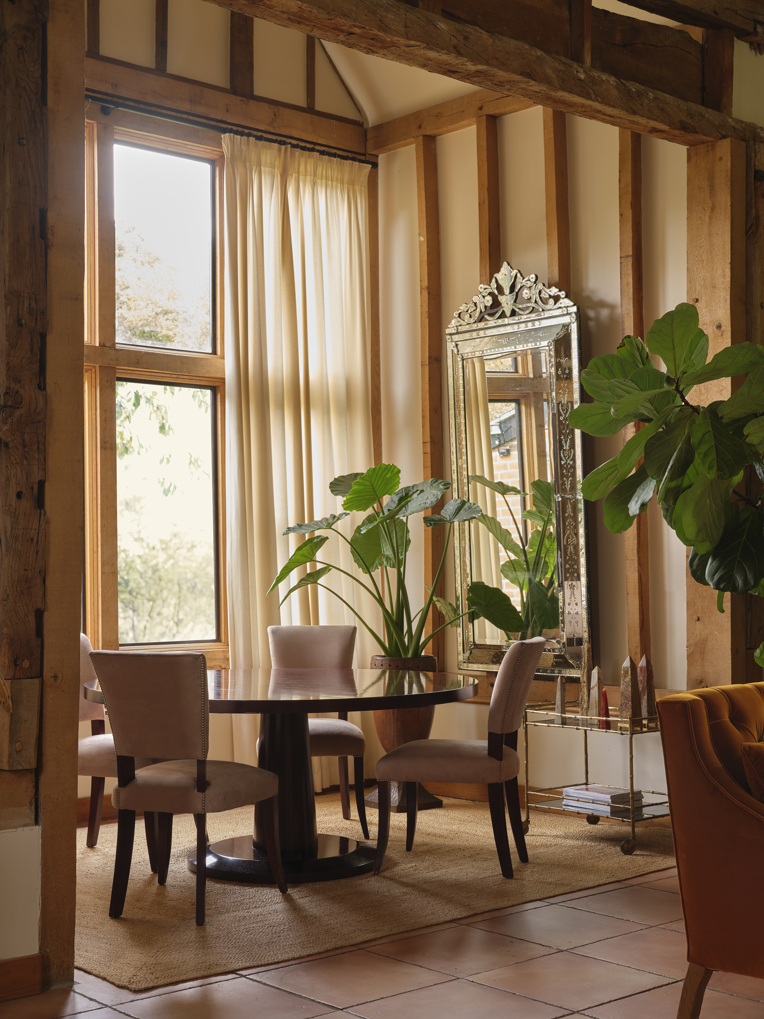 Dining area with a round table, four velvet chairs, potted plants, an ornate mirror, and a brass bar cart.