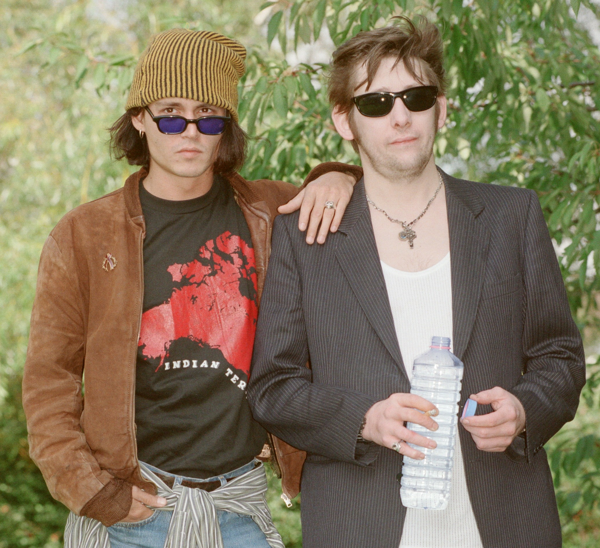Johnny Depp (left) wearing a striped beanie and Shane MacGowan (right) wearing sunglasses, standing together outdoors.