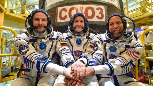 Two cosmonauts and an astronaut wearing spacesuits place their hands on top of each other as they pose for a photograph