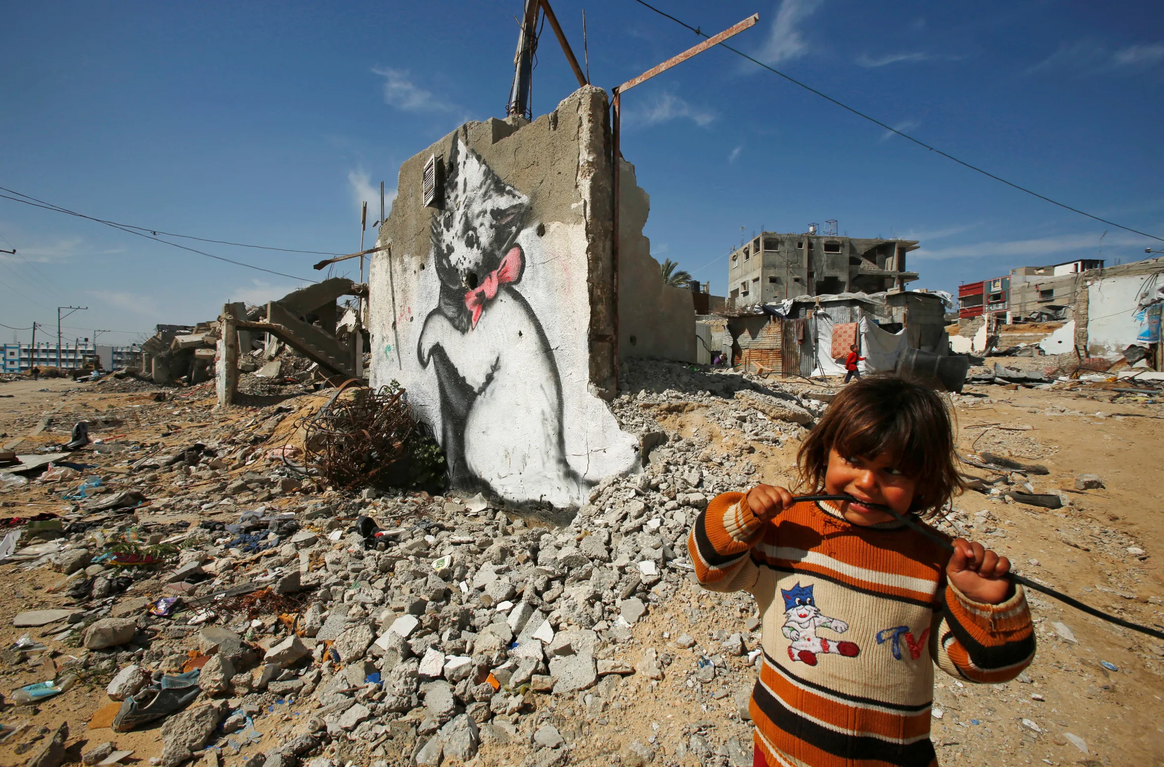 A young Palestinian girl in an orange sweater looks at the camera while chewing on a black cable, standing amidst rubble with a Banksy kitten mural on a destroyed wall in the background.