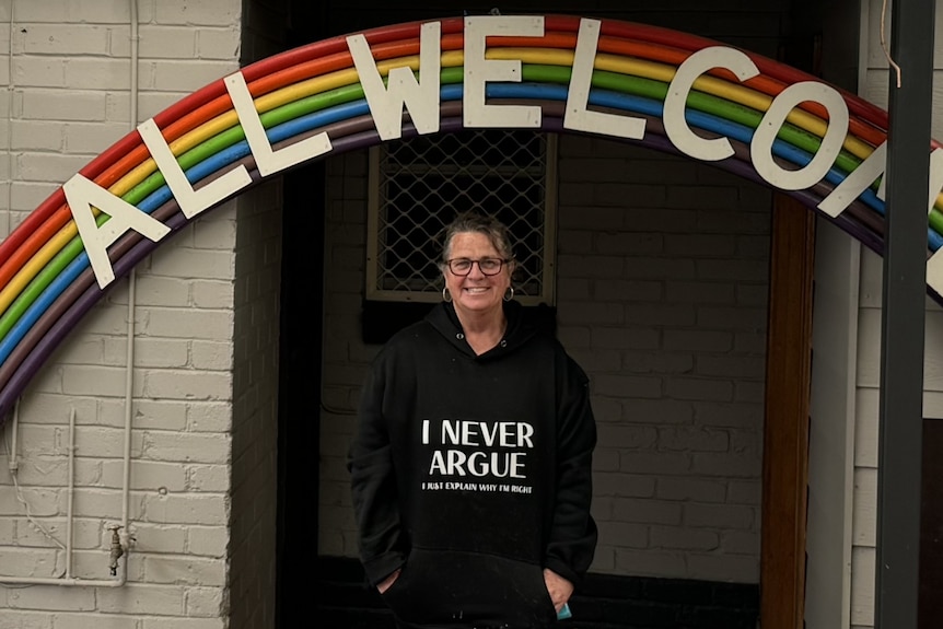 A smiling woman standing under a rainbow sign that says "All welcome".
