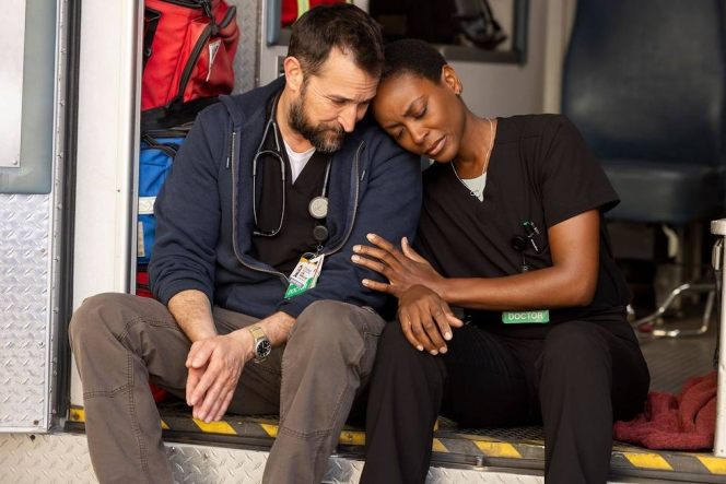 Two doctors in scrubs and stethoscopes, a man and a woman, sit on the back of an ambulance looking weary.
