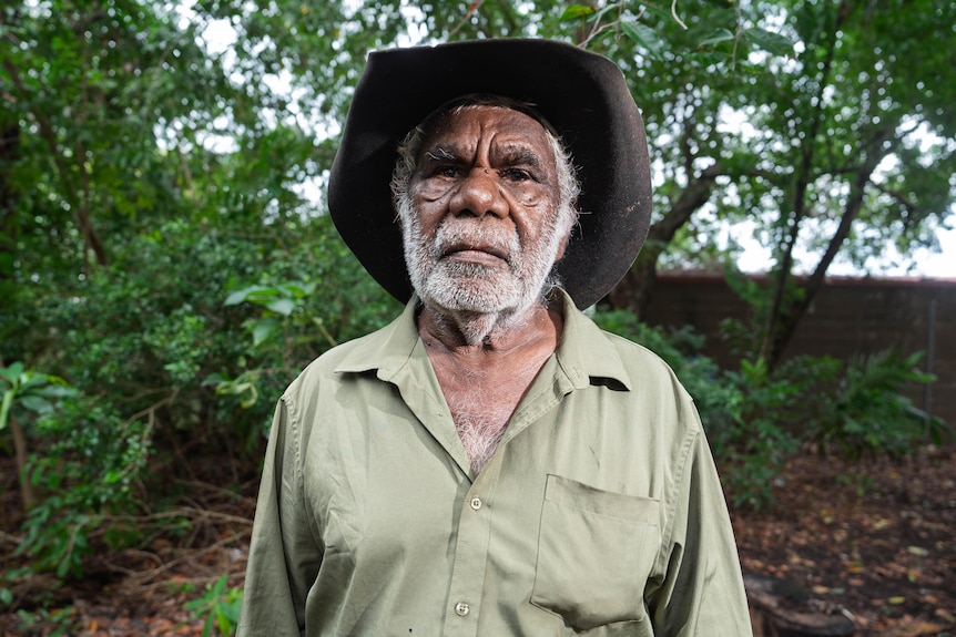 Aboriginal man, gray hair and all around beard, black wide brim hat, khaki button up shirt, serious expression.