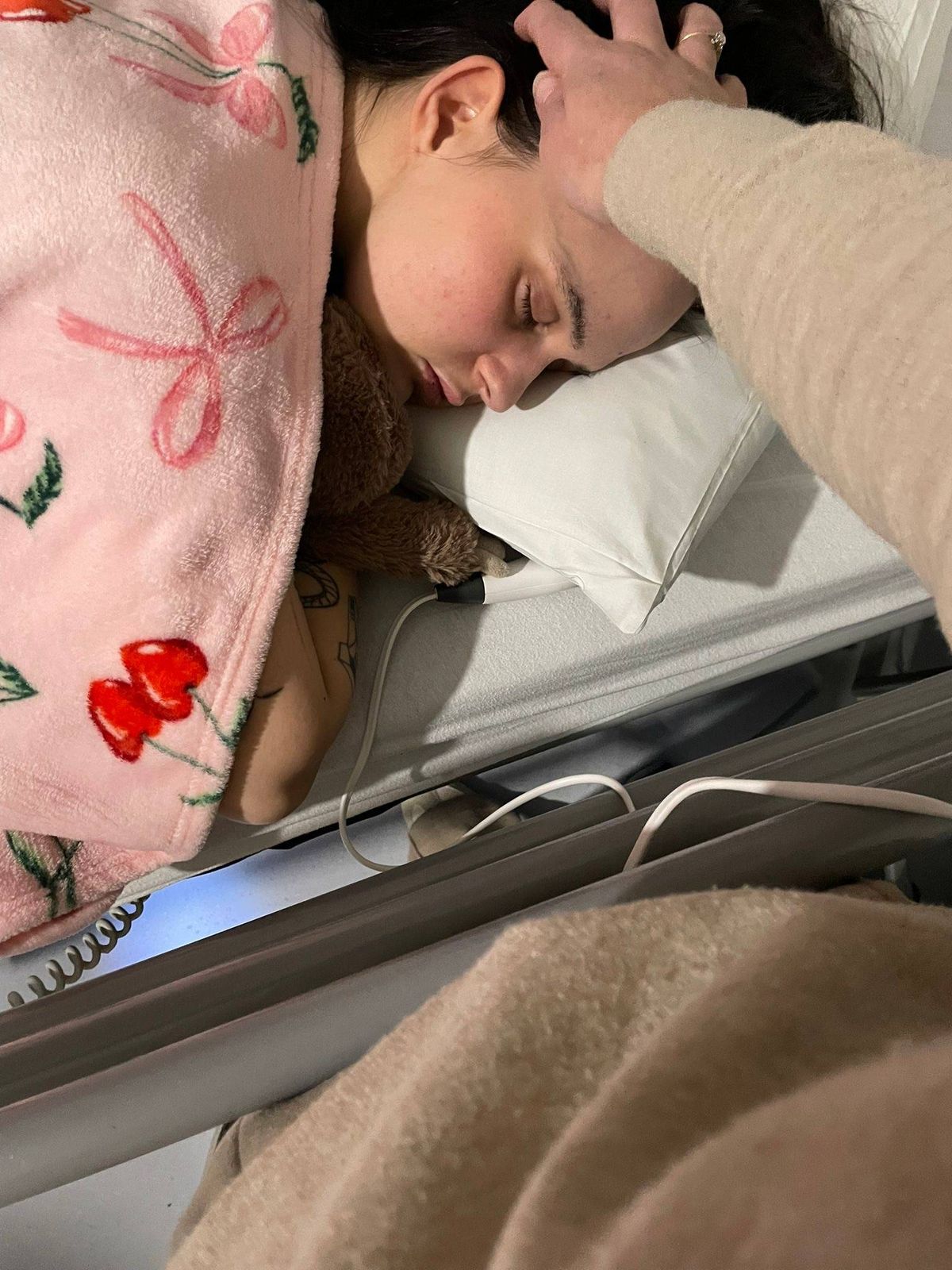 An individual resting on a bed with a pillow, eyes closed, appears to be sleeping. Another person is gently touching the forehead of the sleeping individual with their hand. The person is wearing a light-colored long-sleeved shirt. The sleeping individual is dressed in a pink garment with a floral design.