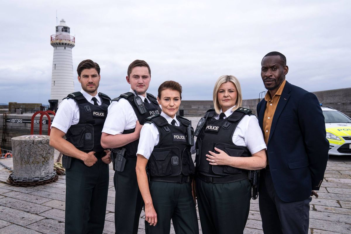 A group of individuals, likely representing a security or enforcement team, are standing together in an outdoor setting. They are dressed in matching uniforms and protective vests, suggesting a professional context. The composition of the group includes both male and female members, and they are standing in close proximity, possibly posing for a photograph. The setting appears to be a well-lit, outdoor environment with a clear sky, contributing to a formal and professional atmosphere.