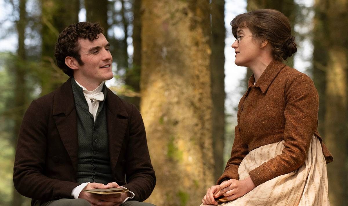 Two individuals, one dressed in formal attire and the other in a period costume, are seated in a forested area, engaged in a conversation while looking at each other.