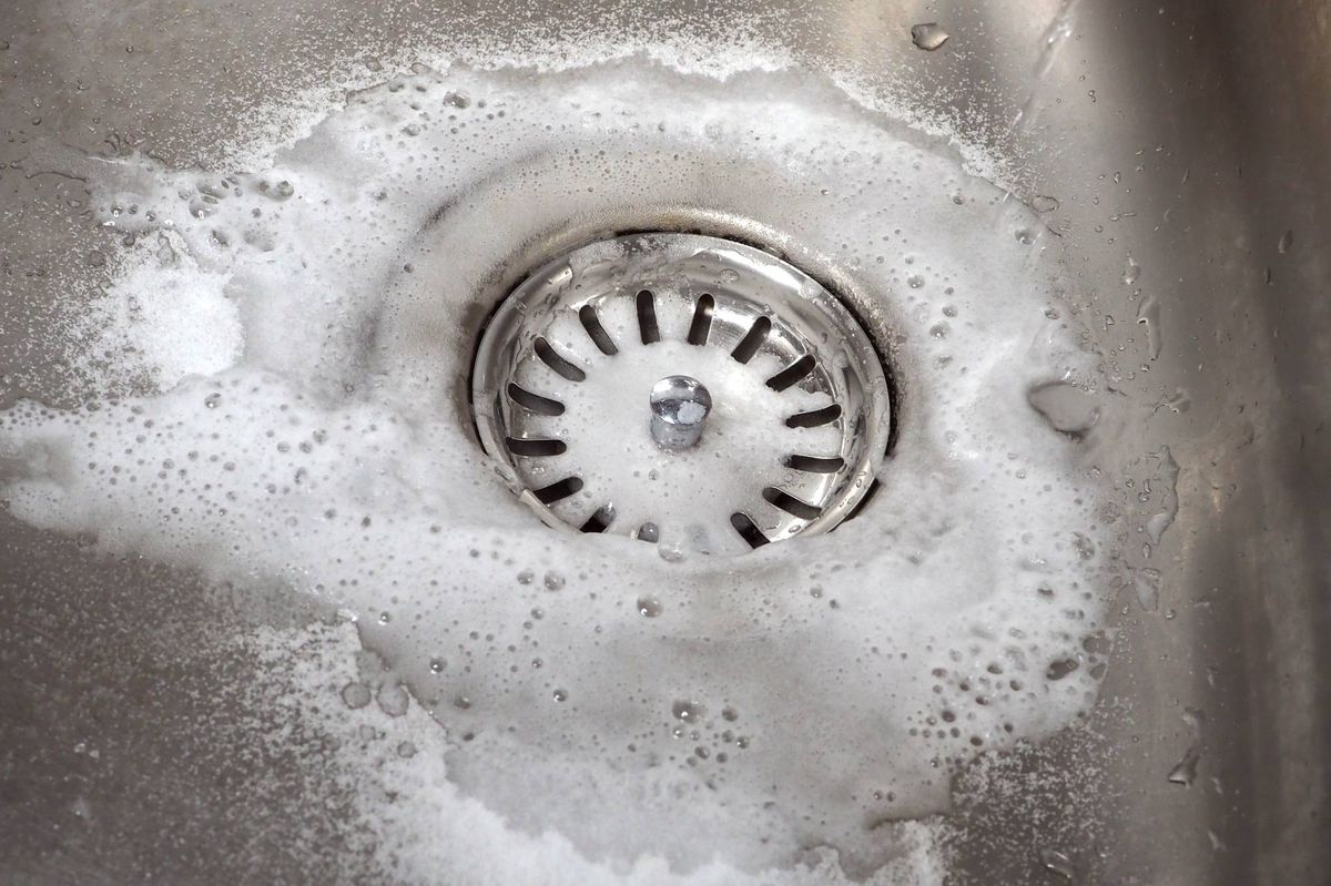 Sink drain cleaning process using baking soda and white vinegar. Close-up of a stainless steel kitchen sink. Tips for home eco-cleaning.