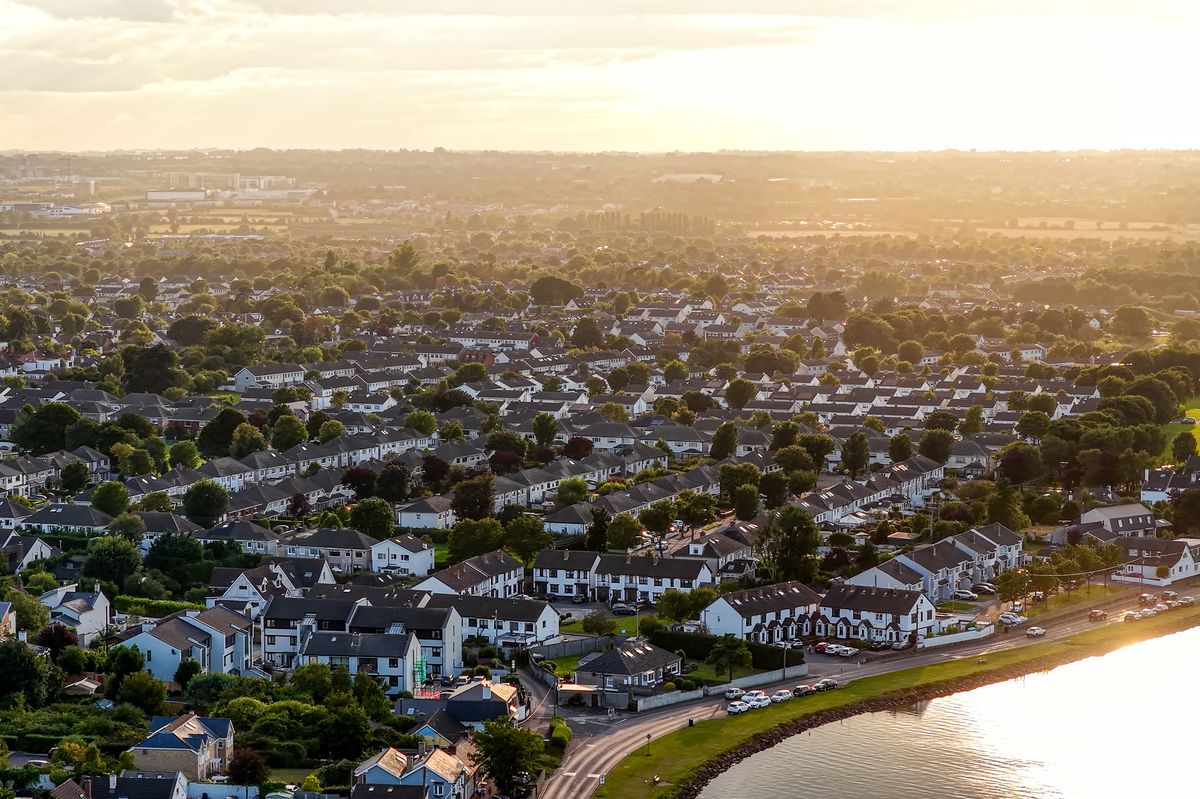 This image captures a scenic aerial perspective of Malahide, a coastal suburban town in north Dublin, Ireland, during the golden hour. The view highlights the harmonious layout of residential housing with distinctive pitched roofs, a marina filled with moored boats, and the calm waters of the estuary. Malahide is known for its historic charm and affluent neighborhoods, with well-preserved urban planning visible in the symmetry and design of the homes. The tranquil coastline and organized urban layout suggest a peaceful and well-developed community, ideal for illustrating real estate, lifestyle, travel, or suburban development themes.