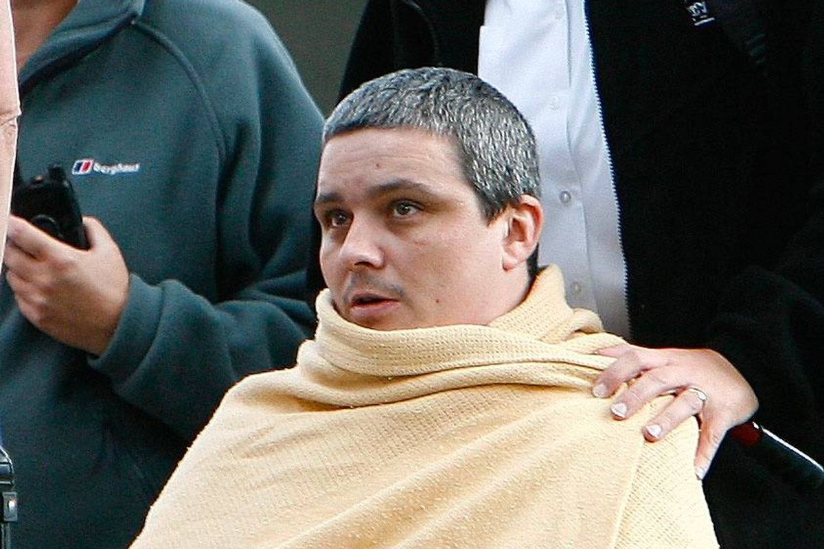 An individual draped in a golden shawl, with another person standing behind them, appears to be engaged in conversation while a third person holds a smartphone in the background.