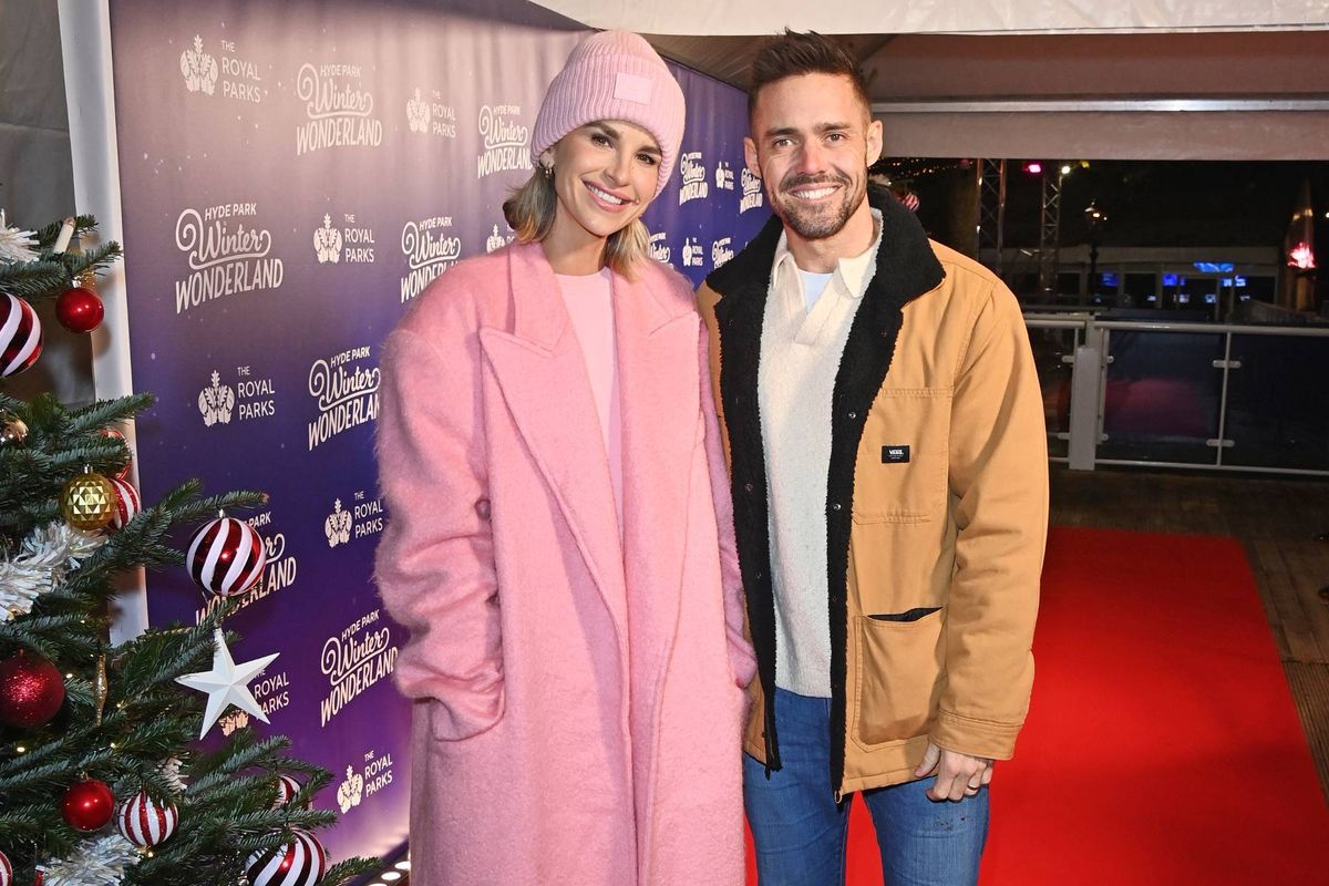 An individual in a pink coat and a person in a brown jacket are standing together on a red carpet, with a festively decorated Christmas tree in the background.
