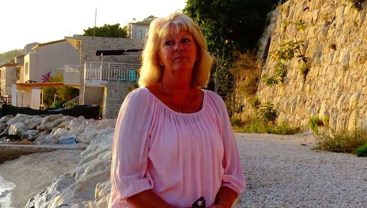 An adult female with blonde hair stands on a gravel path with a stone wall in the background. She is dressed in a pink blouse and appears to be posing for a photograph.