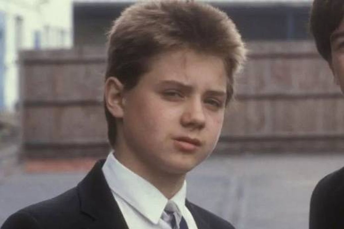 An individual dressed formally in a suit and tie stands in an outdoor setting, gazing to the side while a similarly dressed person stands partially visible behind them. The background features a structured architectural element and an open urban space.