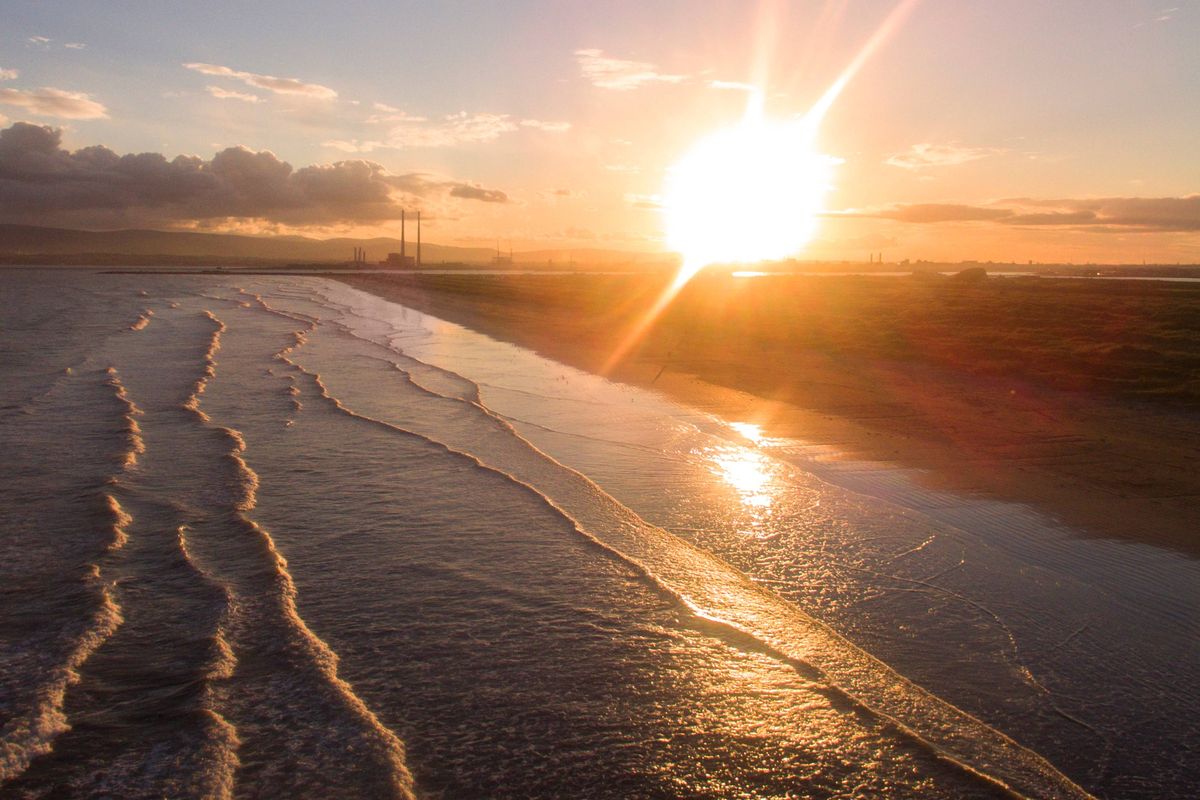 An aerial view captures the majestic scene of the sun setting over a serene body of water, with golden rays reflecting off the surface and creating a shimmering path towards the horizon. The tranquil sky is adorned with soft, wispy clouds, while the distant landmass forms a gentle silhouette against the fading light.
