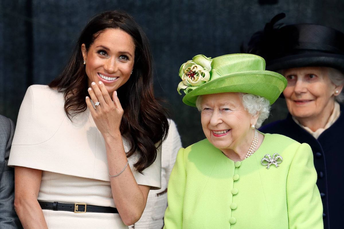 Two individuals, one donning a white dress and the other adorned in a green ensemble with a matching hat, are standing close to each other, with the person in the white dress appearing to be engaged in a conversation.