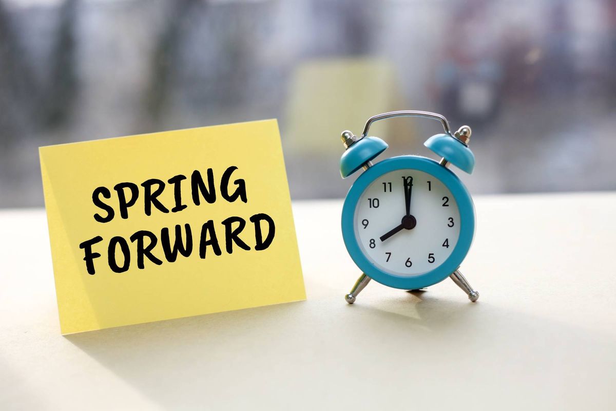 An image features a small blue alarm clock next to a yellow sticky note with the words "Spring Forward" written on it. The alarm clock shows the time as 11:50.