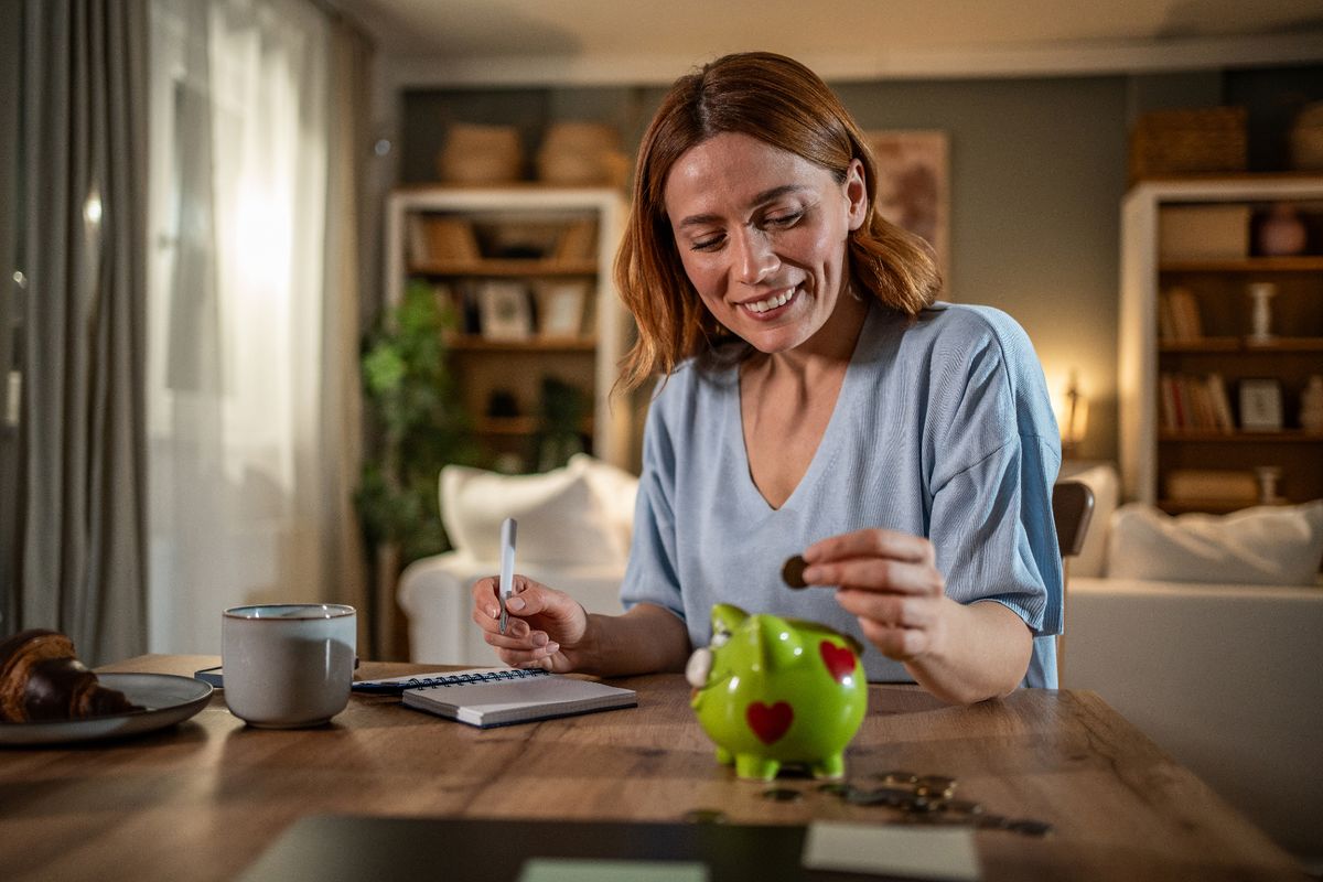 Smiling woman placing coins into a piggy bank while jotting down notes about home finances, actively planning her budget and focusing on saving money for future goals