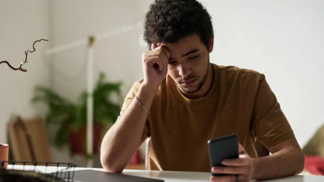 A young man looks at a phone with his hand on his head as he sits in front of a computer