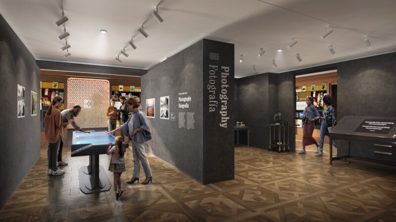 A modern museum exhibit features people interacting with touchscreens and displays about photography. The room has dark walls, wood floors, and informational panels, with adults and children exploring the space.