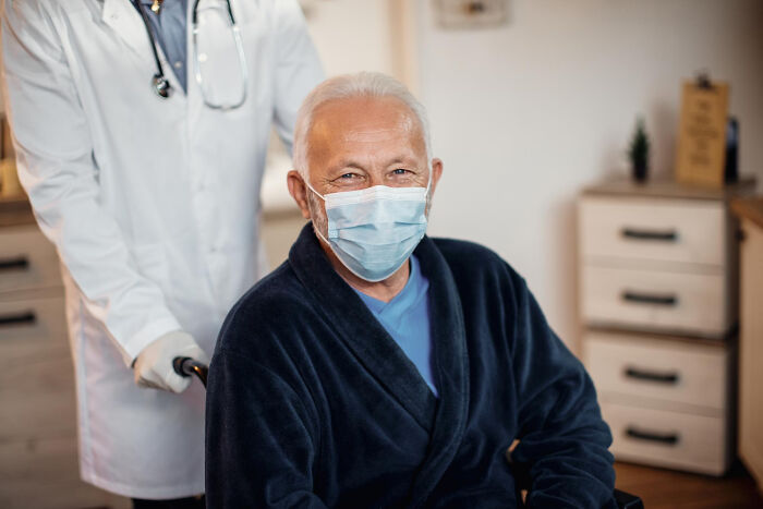 Elderly man in a mask sitting in a wheelchair with a doctor behind him, illustrating patients faking symptoms cases.