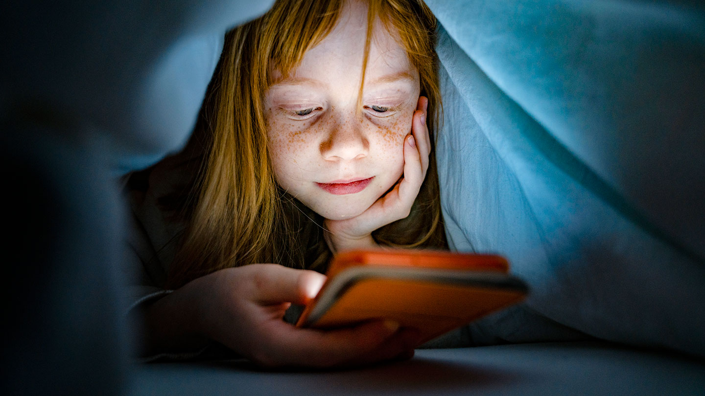a pale child with freckles hides under a blanket to use a cell phone at night