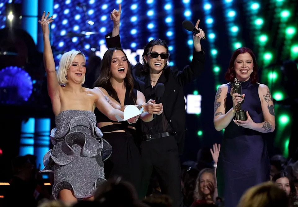 Four people celebrate on stage at an awards event; one holds a trophy. They are dressed in stylish and diverse outfits, showing excitement and joy