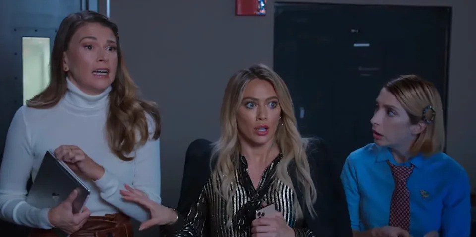 Three women look surprised in an indoor setting. One holds a tablet, another a phone, and the third wears a ribbon-tied shirt