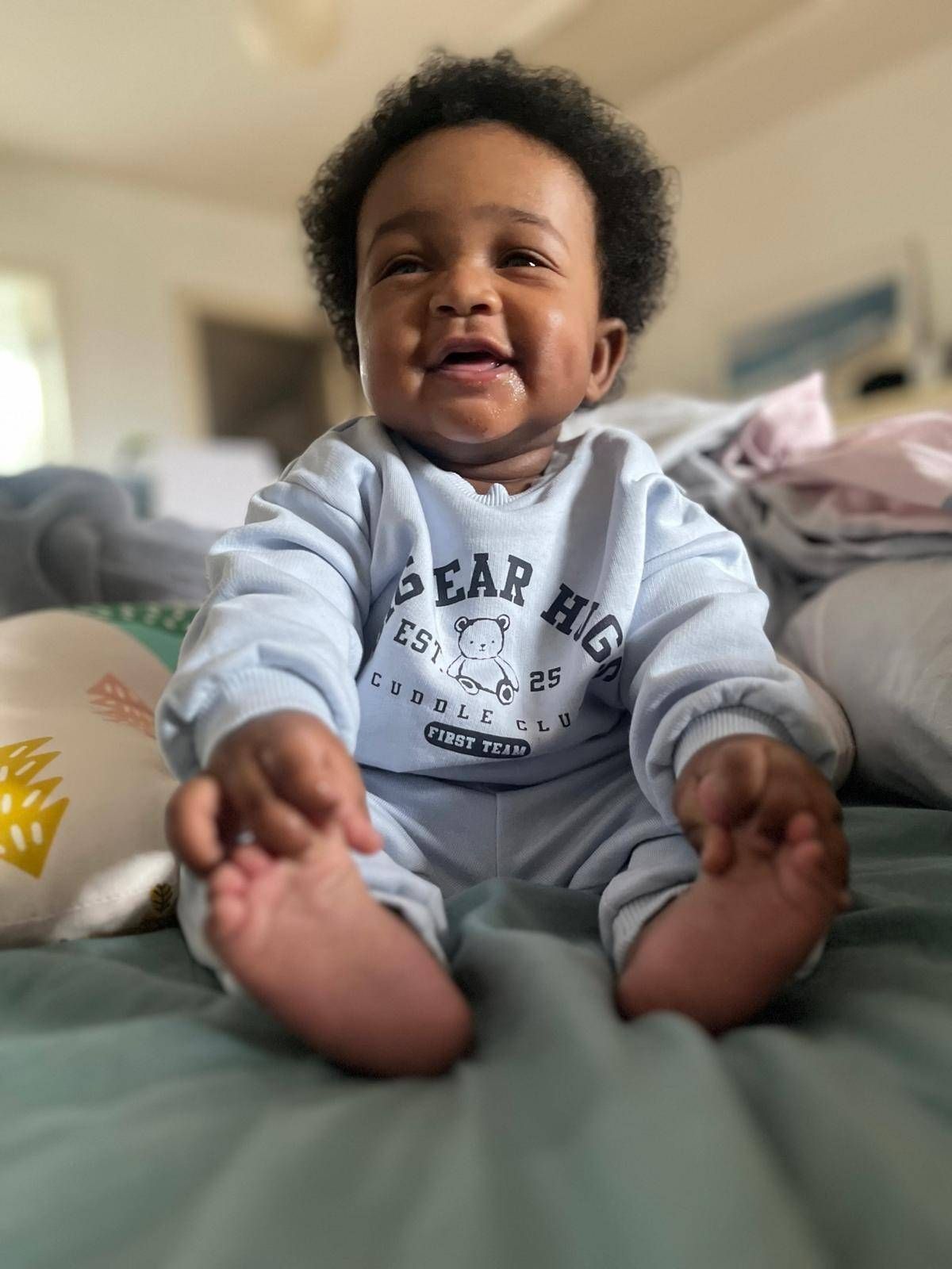 An image depicts a young child, possibly a toddler, seated in a domestic environment. The child is dressed in a light-colored sweatshirt featuring printed text, and the expression on their face suggests a moment of joy or amusement. The background includes elements of a living space, with various furnishings and decorative items visible.