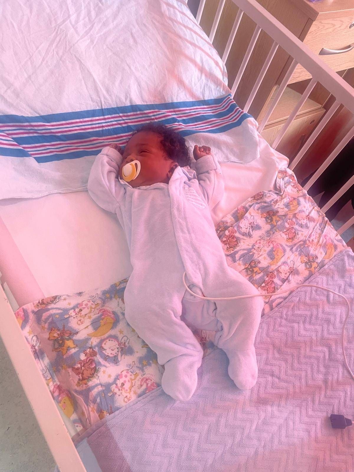 A newborn baby is resting on a bed, clad in a pink onesie and holding a pacifier, lying on a bed adorned with a patterned blanket and pillows.