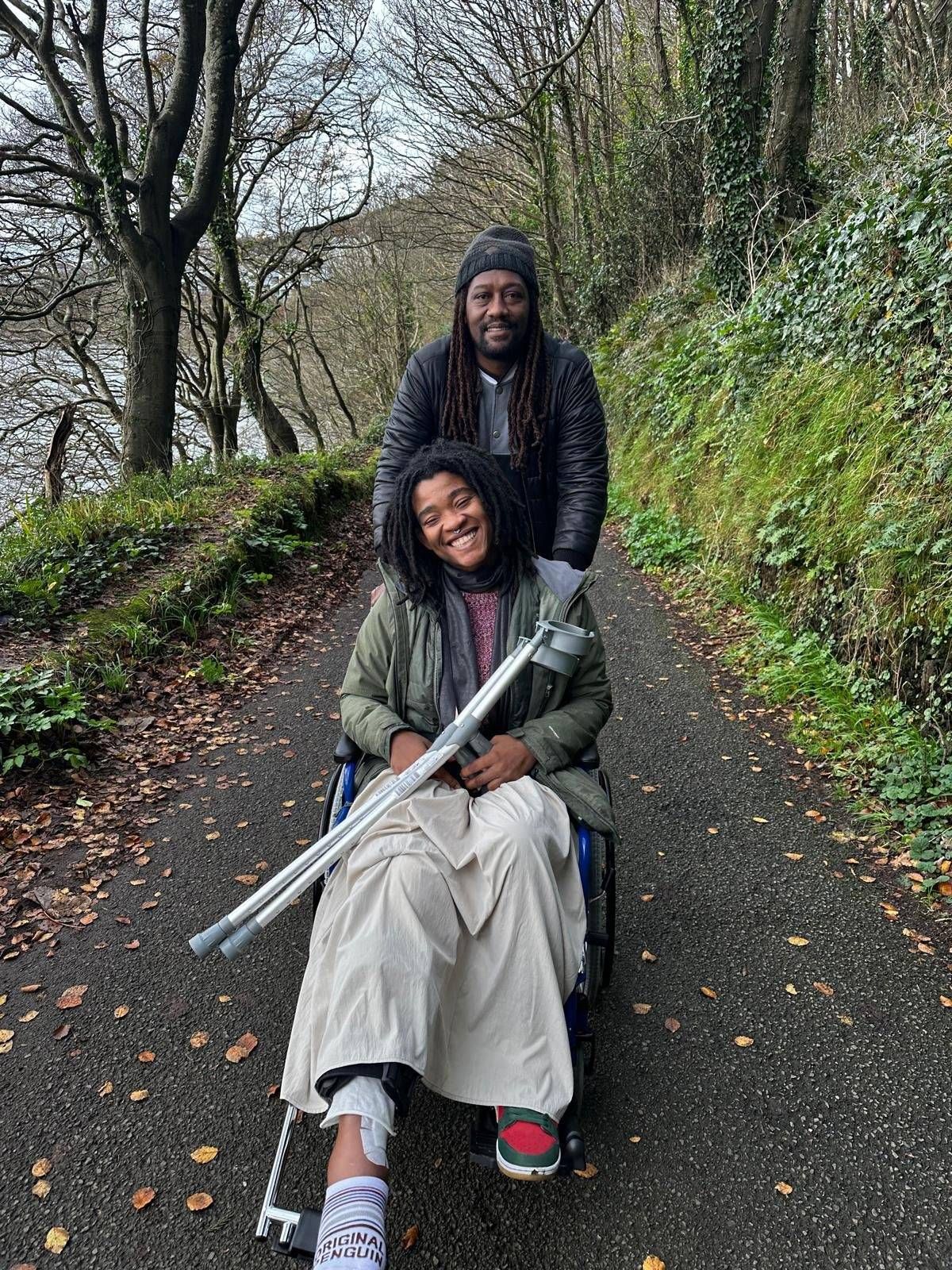 An individual seated in a wheelchair is positioned on a paved pathway, with another person standing behind them. The person in the wheelchair is holding a pair of long, metallic rods. The pathway is flanked by lush greenery and vegetation, with a water body visible on the left side.