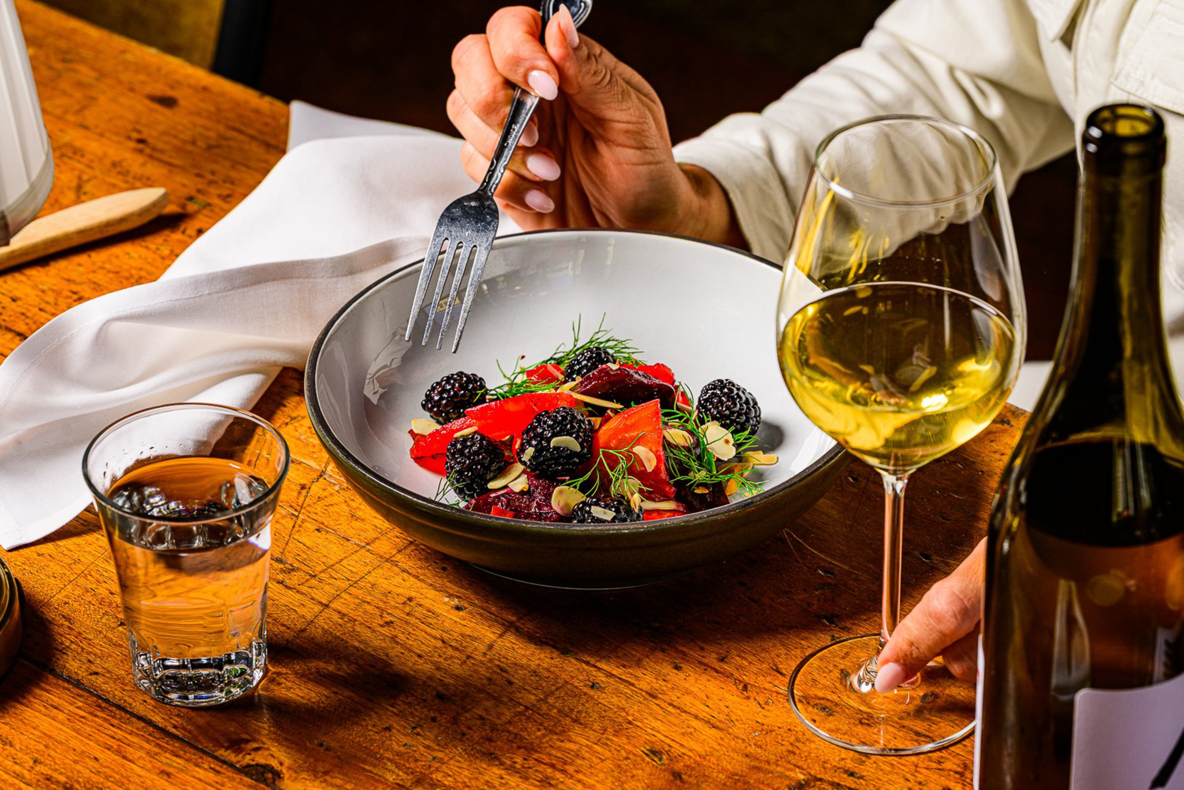 A hand holds a fork over a bowl of salad with blackberries, red vegetables, and greens, next to a glass of white wine and a bottle on a wooden table.