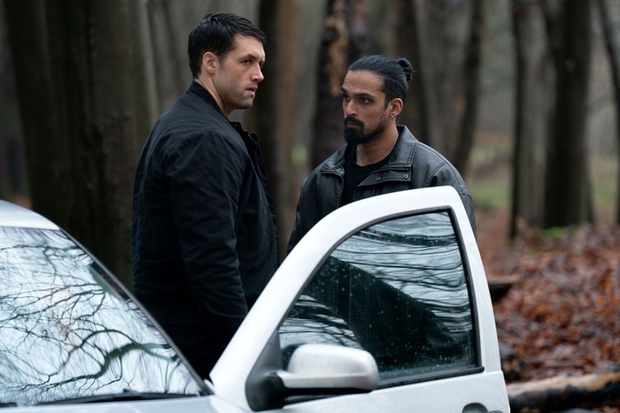 Ravi and Mark stood amid some trees in front of an open car door in a scene from EastEnders