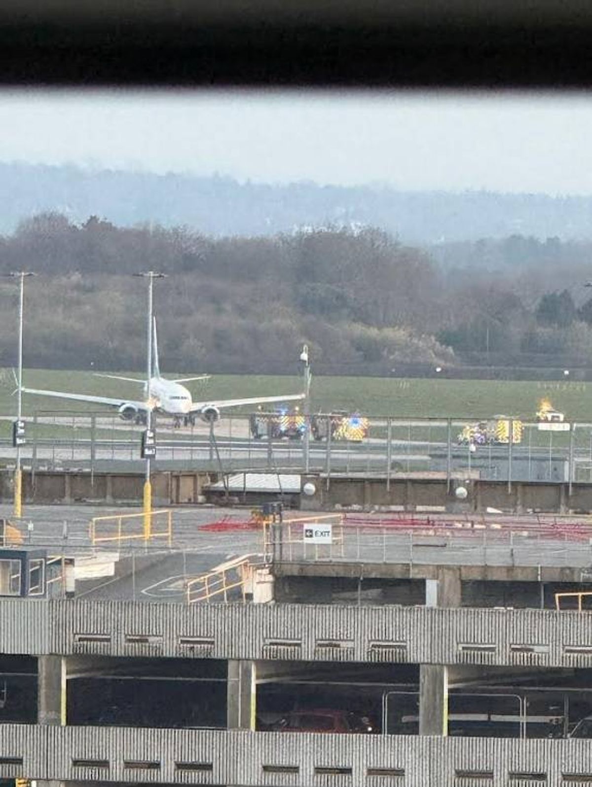 An aerial view of an airport runway features an airplane positioned on the tarmac. The runway is flanked by a series of poles and traffic signs. In the background, a hill with trees is visible against a hazy sky.