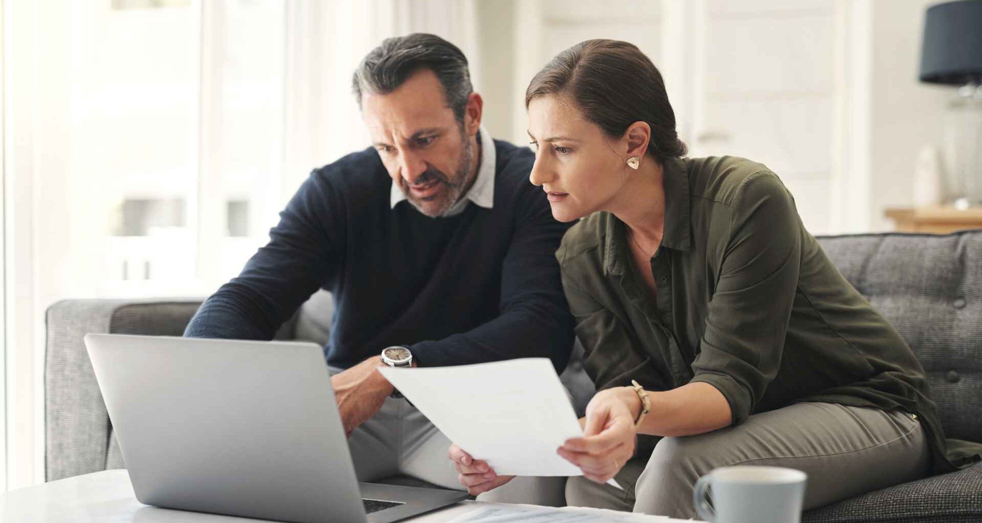 A couple reviews financial paperwork together while sitting at a table with a laptop open in front of them.
