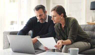 A couple reviews financial paperwork together while sitting at a table with a laptop open in front of them.