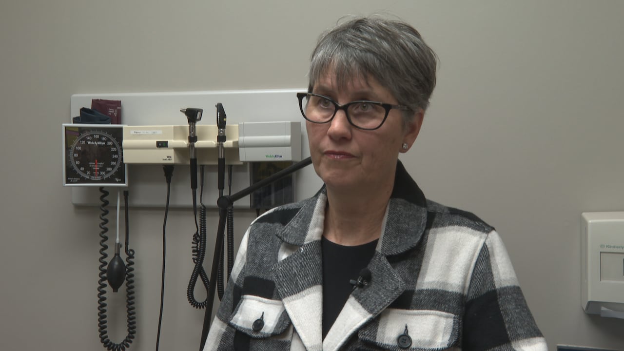 A woman with short hair and glasses sits in front of a wall with medical equipment behind her