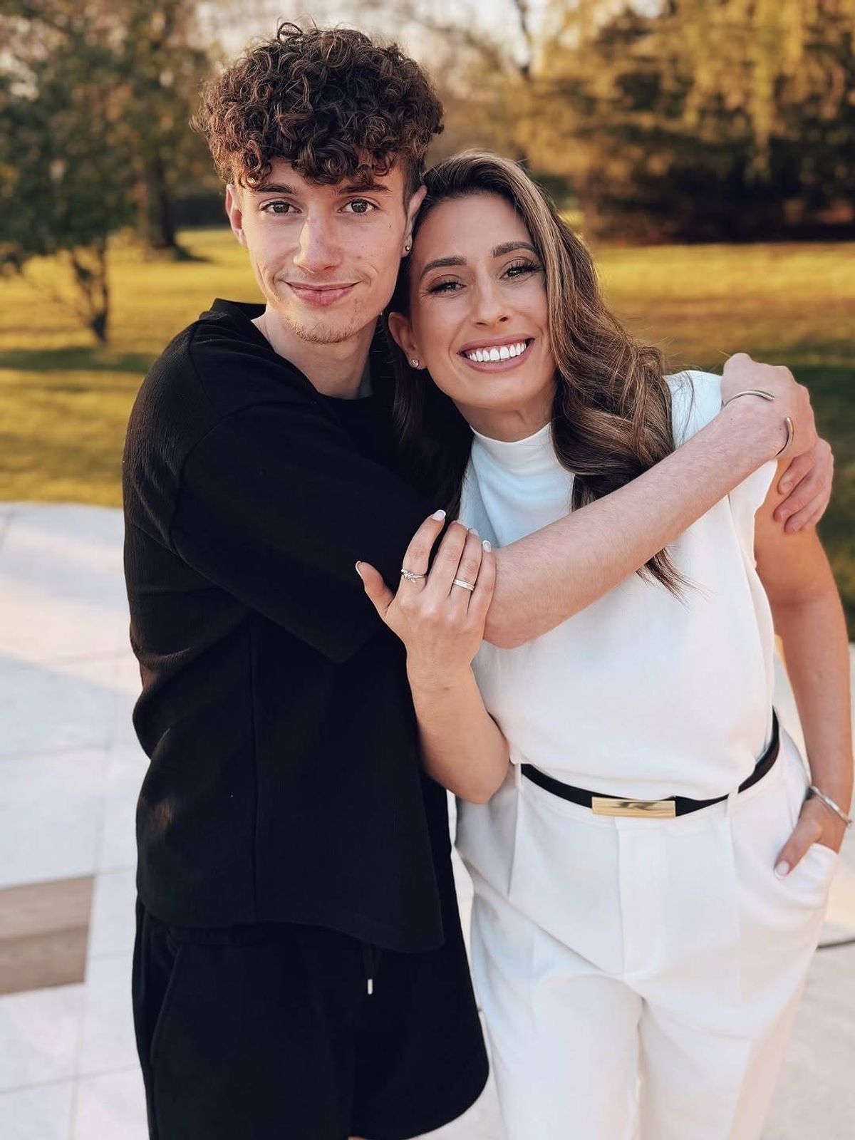A man and a woman standing together in an outdoor setting, smiling and embracing each other while surrounded by a lush green landscape with trees and grass.