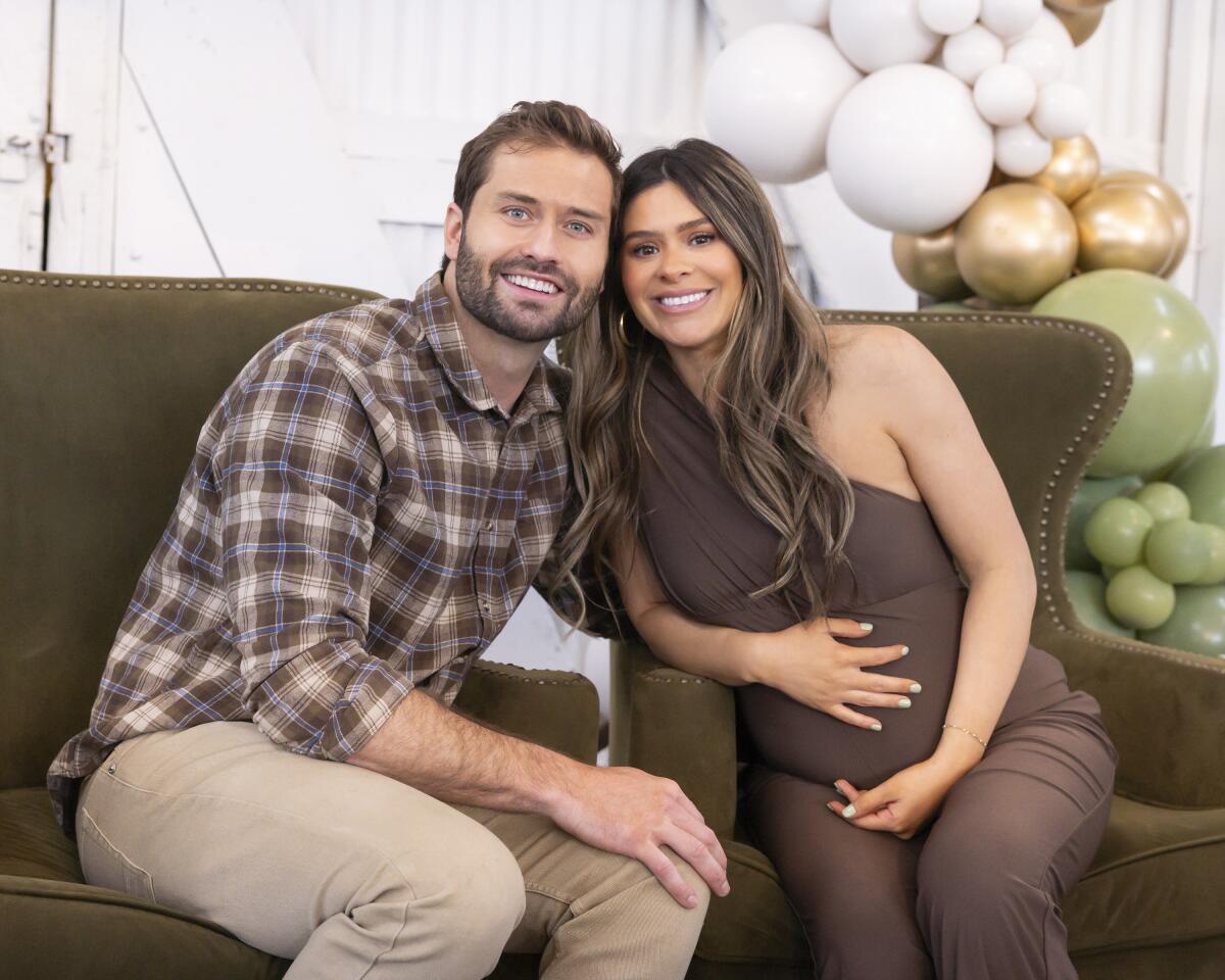 A man in a plaid shirt and a pregnant woman in a brown jumpsuit sit on a couch smiling and leaning their heads together.