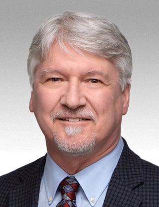 A man with grey hair and a short grey goatee is wearing a checkered jacket and red and blue tie.