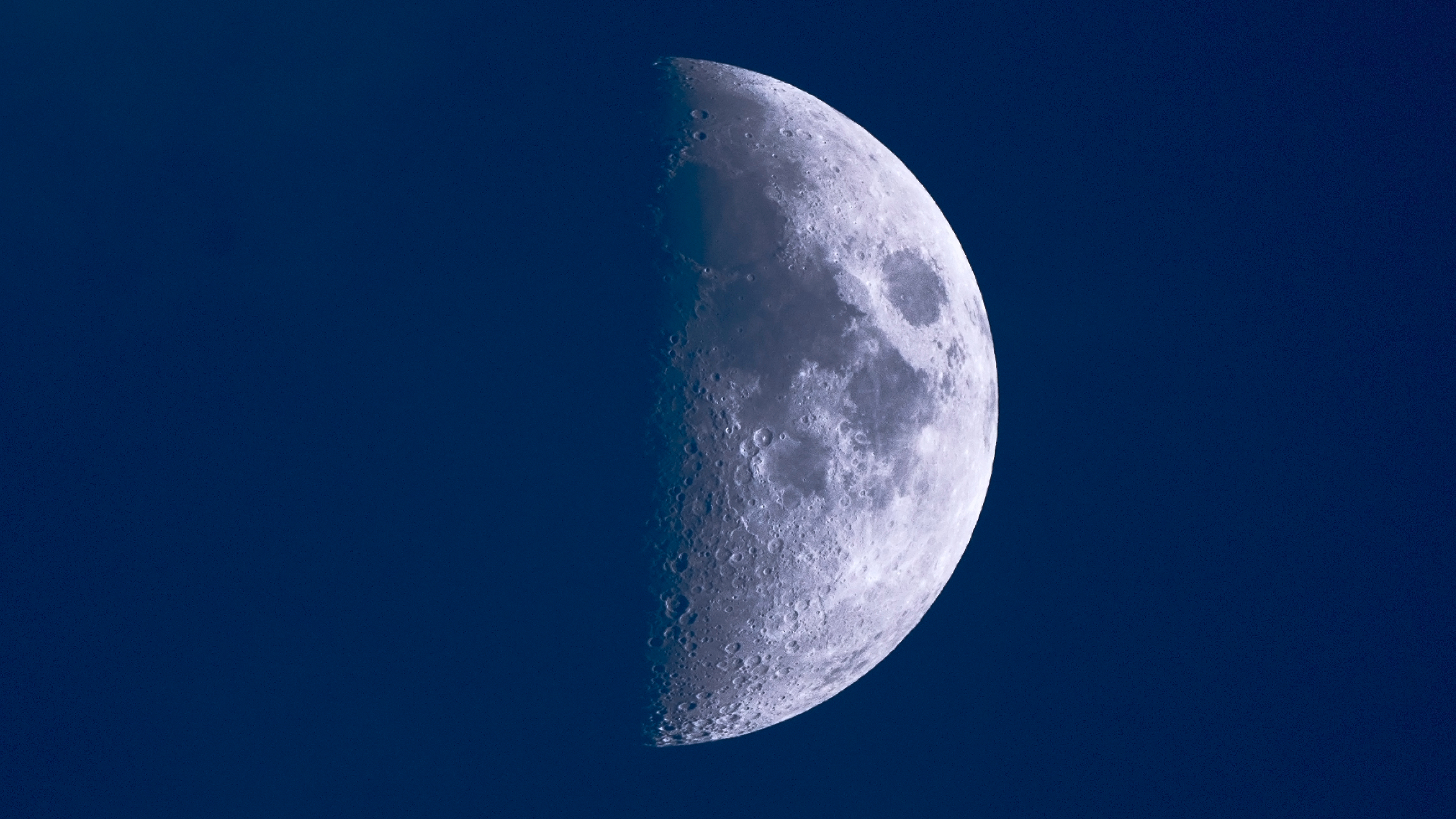 The moon is shown in a dark blue sky with its right half lit and its left loft to shadow. Ancient dark lava fields known as lunar maria cover swathes of its surface.