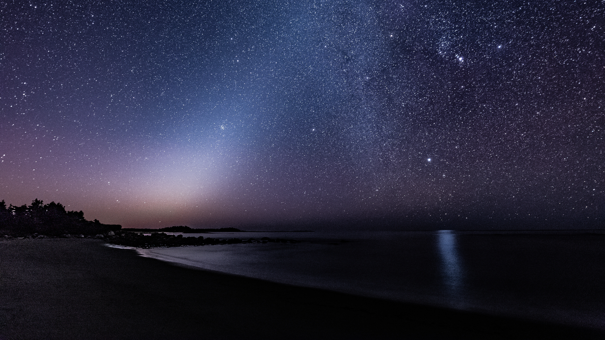 A photograph of the night sky showing the hazy glow of zodiacal light shining to the left of the glowing band of the Milky Way behind the shoreline of a lake